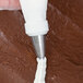 A person using an Ateco drop flower piping tip with a pastry bag to pipe white frosting on a brown surface.
