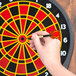 A hand holding a red and black Arachnid SFR150 soft tip dart in front of a dart board.
