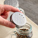A hand is holding a metal shaker lid with multiple holes above a glass jar containing ground spices.