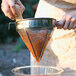 A person pouring orange liquid through a stainless steel China cap strainer.