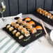 A black Acopa stoneware plate with sushi on a black table.