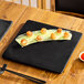 A large, square matte black stoneware flat plate displayed on a wooden table with food arranged on top.