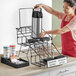 A woman in an apron putting a coffee thermos into a black wire airpot rack.