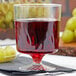 A clear plastic wine goblet filled with red liquid next to grapes on a table.