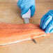 A gloved hand uses a Choice curved boning knife to cut a piece of salmon on a cutting board.