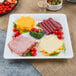 A Fineline white plastic square cater tray with cheese, meat, and vegetables.