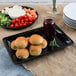 A black Fineline rectangular cater tray with rolls and vegetables and a jar of jam on it.
