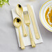 A Visions Heavy Weight Elegant gold plastic cutlery set with a spoon, fork, and knife on a napkin next to a bowl of soup.