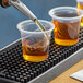 A person pouring brown liquid from a pipette into clear plastic shot glasses on a tray.