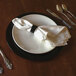 A Tabletop Classics black plastic charger plate with a beaded rim with silverware and a white napkin on it.