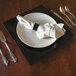 A black Tabletop Classics by Walco plastic charger plate on a table with a white plate on top and silverware