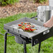 A person using a Backyard Pro stainless steel griddle plate with handles to cook food on a grill.
