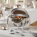 A round stainless steel chafing dish with a roll-top lid is displayed on a white tablecloth, containing cooked food and accompanied by serving utensils and plates.