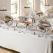A buffet table set with shiny stainless steel chafing dishes containing various hot foods, with serving utensils and small label cards in front of each dish.