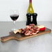 An American Metalcraft carbonized bamboo serving peel with meat, grapes, and cheese on a counter with a glass of wine.