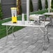A Regency stainless steel folding work table on a patio with a grill and a silver metal container on it.