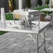 A Regency stainless steel folding work table on a sidewalk with a chafing dish on it.