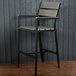 A BFM Seating black and gray aluminum outdoor bar stool with a synthetic teak back and seat leaning against a wall.