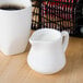 A Tuxton white china creamer pitcher on a table with a white mug of coffee.