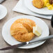 A Tuxton TuxTrendz bright white china plate with a croissant and butter on it with a knife.