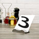 A Tablecraft stainless steel wavy table number holder on a table with wine bottles.