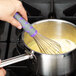 A hand with a grey and purple handle using a Vollrath Jacob's Pride stainless steel whisk to mix liquid in a pot.