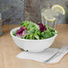 A white bowl of salad with lettuce on a table with a glass of water and a lemon wedge.