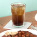 A Libbey Gibraltar beverage glass filled with brown liquid and ice on a table with a plate of food.