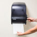 A hand pulling a black paper towel from a San Jamar Classic Lever Roll Towel Dispenser.