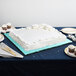 A white square cake on a blue Enjay cake board on a table with a fork and plates.