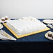 A white table with a white and gold frosted cake on a gold square cake board.
