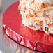 A close up of a piece of cake on a red Enjay round cake board with white frosting and coconut flakes.