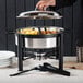A person using an Acopa wrought iron chafer stand to serve food on a table outdoors.