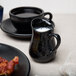 An American Metalcraft black porcelain bell creamer on a table with a cup of white liquid and a plate of bacon.