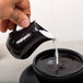 A hand pouring milk from an American Metalcraft black porcelain bell creamer into a cup of coffee.