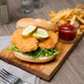 A chicken sandwich with pickles and a glass of ketchup on a wooden board.
