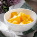 A white bowl of cubed yellow mango on a table with a spoon and a napkin.