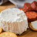 A round white Boursin cheese on a wooden board with crackers and pepperoni.