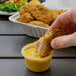 A hand dipping a piece of fried chicken into a small plastic container of Ken's Honey Mustard.