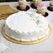 A white table with a white Enjay Fold-Under Gold Round Cake Drum holding a white cake with frosting and cupcakes.