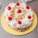 A white frosted cake with red cherries on a gold cake drum.