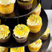 A black Enjay 3-tier cupcake stand with cupcakes frosted in yellow and sprinkles.