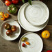 A stack of Oneida Marble porcelain plates with a raised rim on a wood surface with bowls and tomatoes.