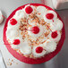 A red Enjay round cake drum under a cake with white frosting and red cherries on top.