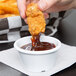 A person dipping a piece of fried chicken into a white plastic ramekin of sauce.