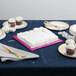 A pink Enjay square cake drum on a table with a white cake and cupcakes.