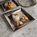 Two American Metalcraft square stainless steel trays with food on them on a marble table.