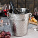 A Tablecraft stainless steel wine bucket with a bottle of wine and two glasses inside.