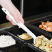 A person holding a white Cambro salad bar spoon over a tray of food.