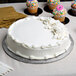 A white frosted cake on a white Enjay round cake drum on a table with cupcakes and a knife.